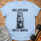 Light blue t-shirt with distressed print of a cat holding a kettlebell, text "BUILT DIFFERENT MOSTLY ANNOYED".
