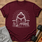 Maroon t-shirt with white graphic of a person in a hood sitting on a bench with a gym bag.