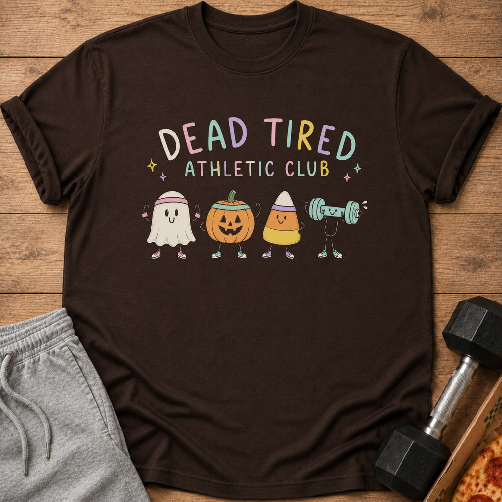 Brown t-shirt featuring a Dead Tired Athletic Club graphic with cartoon Halloween characters, resting on a wooden surface with sweatpants and a dumbbell.