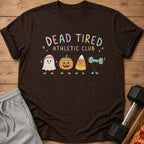 Brown t-shirt featuring a Dead Tired Athletic Club graphic with cartoon Halloween characters, resting on a wooden surface with sweatpants and a dumbbell.