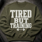 Olive green sweatshirt with distressed white text reading "TIRED BUT TRAINING" and a barbell icon.