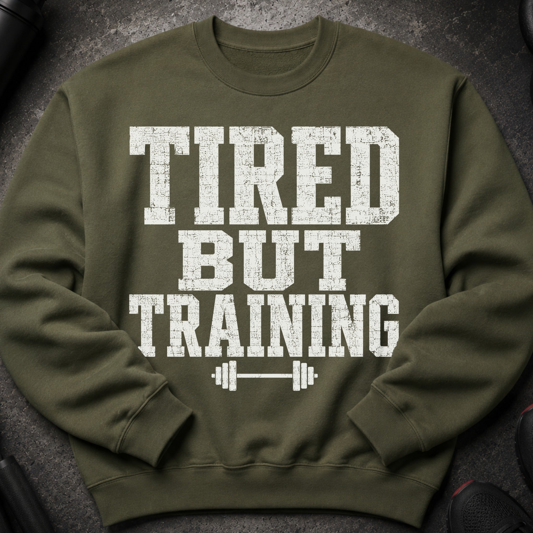 Olive green sweatshirt with distressed white text reading "TIRED BUT TRAINING" and a barbell icon.