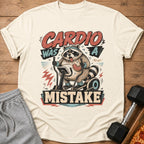 Cream t-shirt with raccoon on elliptical, reads "CARDIO WAS A MISTAKE."