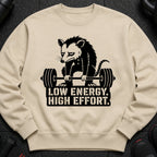 Tan sweatshirt with a black graphic of a sad opossum lifting a barbell and text.