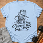 Light blue t-shirt with a distressed drawing of an opossum tangled in resistance bands, text reads "Stretched Thin, Still Here."