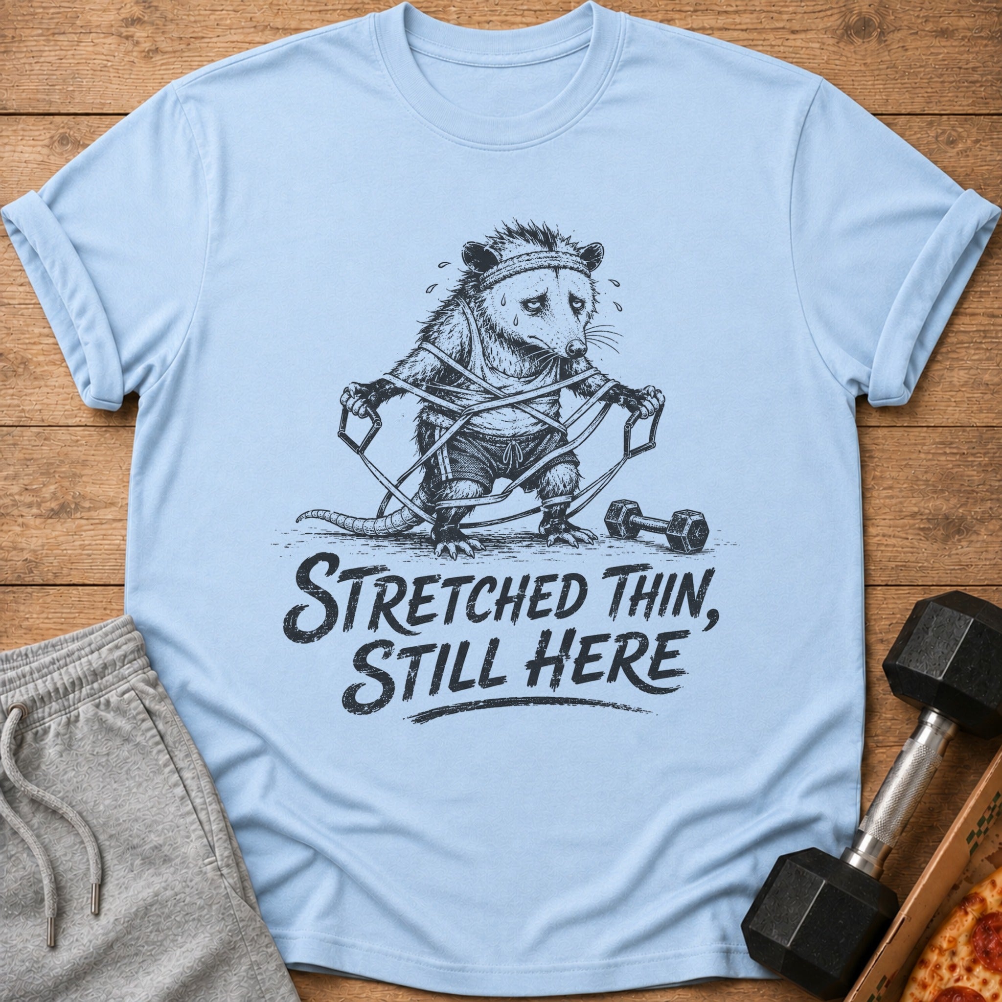 Light blue t-shirt with a distressed drawing of an opossum tangled in resistance bands, text reads "Stretched Thin, Still Here."