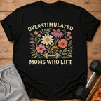 A black t-shirt printed with the text Overstimulated Moms Who Lift, floral graphics, and a dumbbell.
