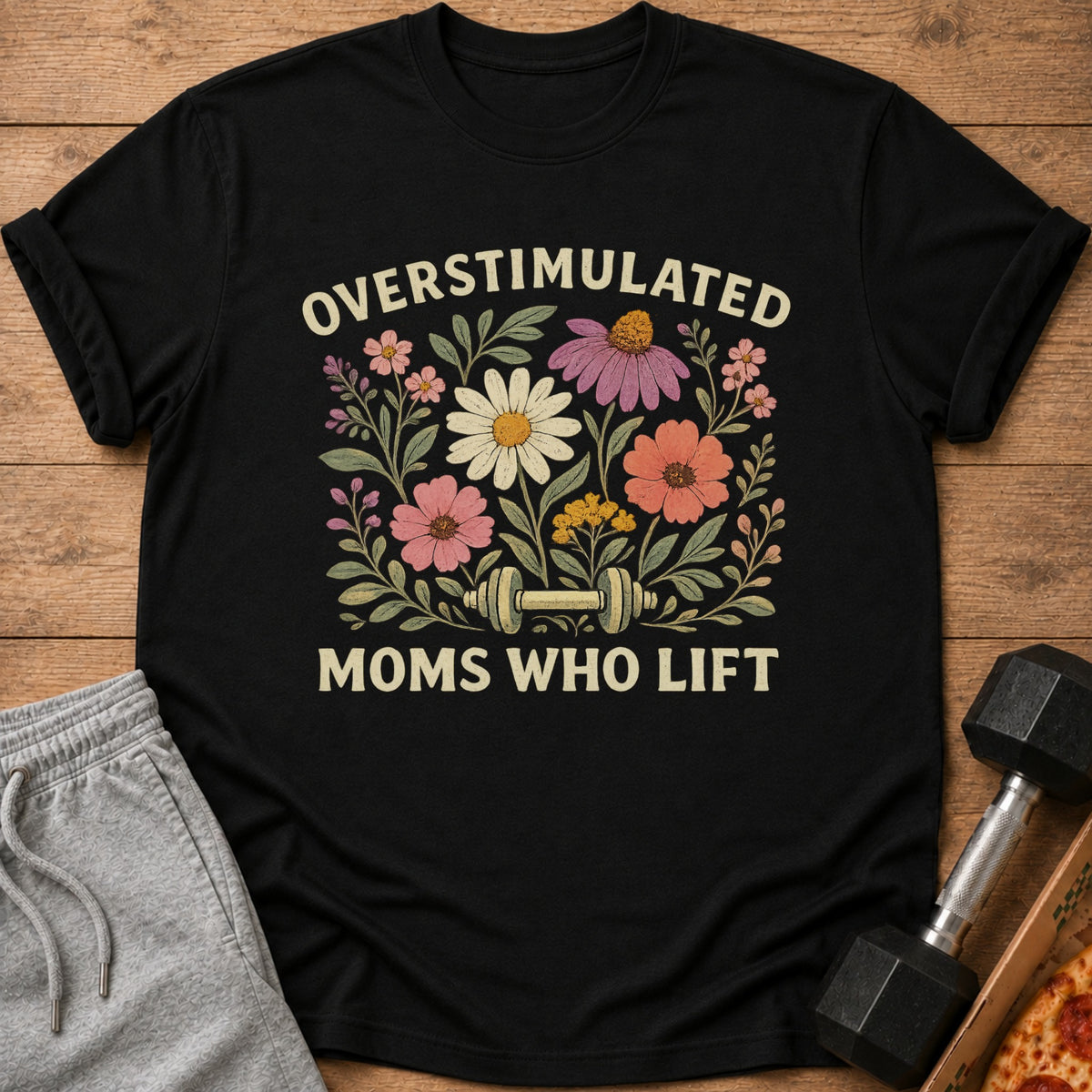 A black t-shirt printed with the text Overstimulated Moms Who Lift, floral graphics, and a dumbbell.