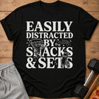 Black t-shirt with "Easily Distracted By Snacks & Sets" text and illustrations.