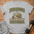 Grey t-shirt with a snail lifting weights and the text "Progress is technically happening."