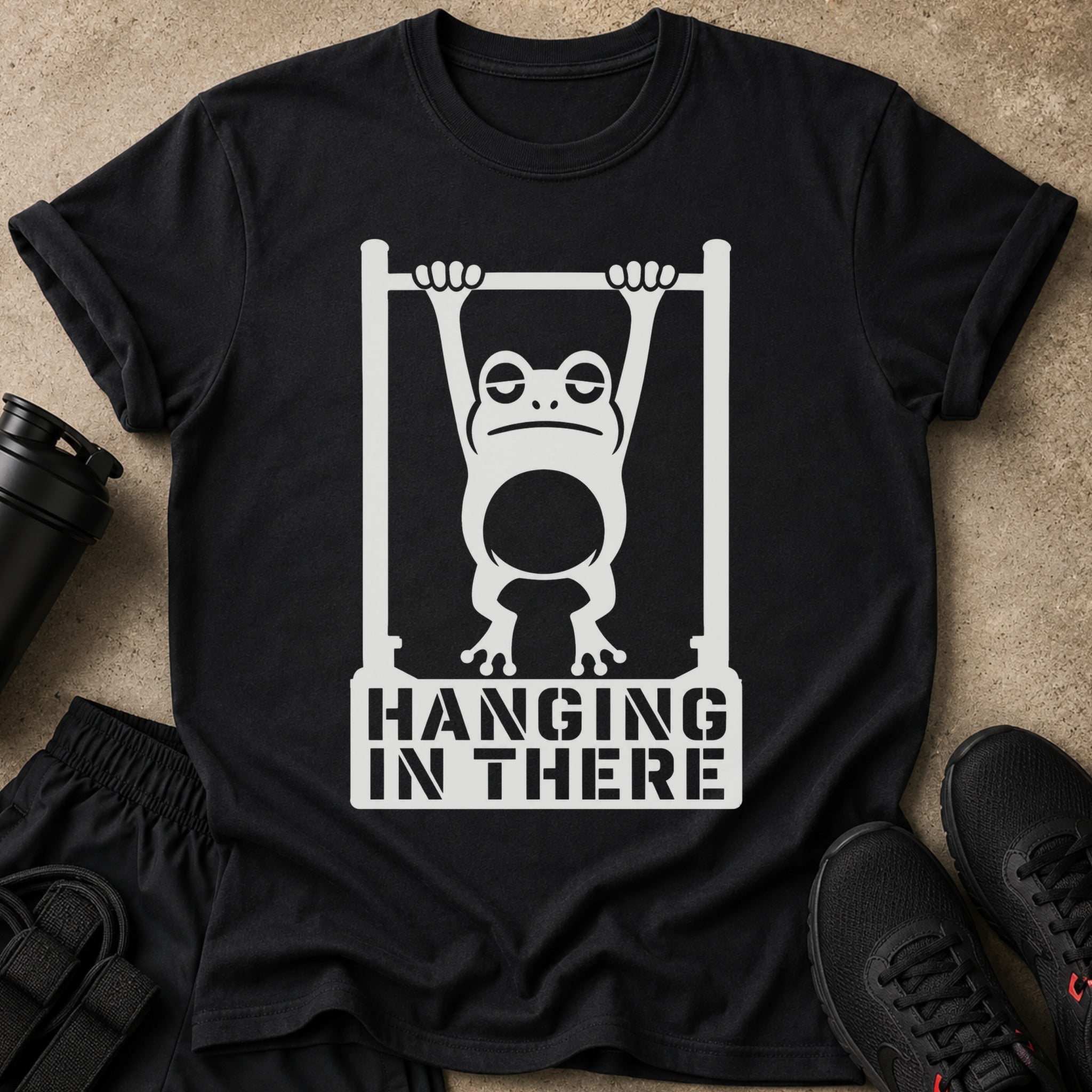 Black t-shirt with white graphic of a tired frog hanging from a bar, text reads "HANGING IN THERE".