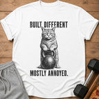 White t-shirt with a cat holding a kettlebell and the text "BUILT DIFFERENT MOSTLY ANNOYED."