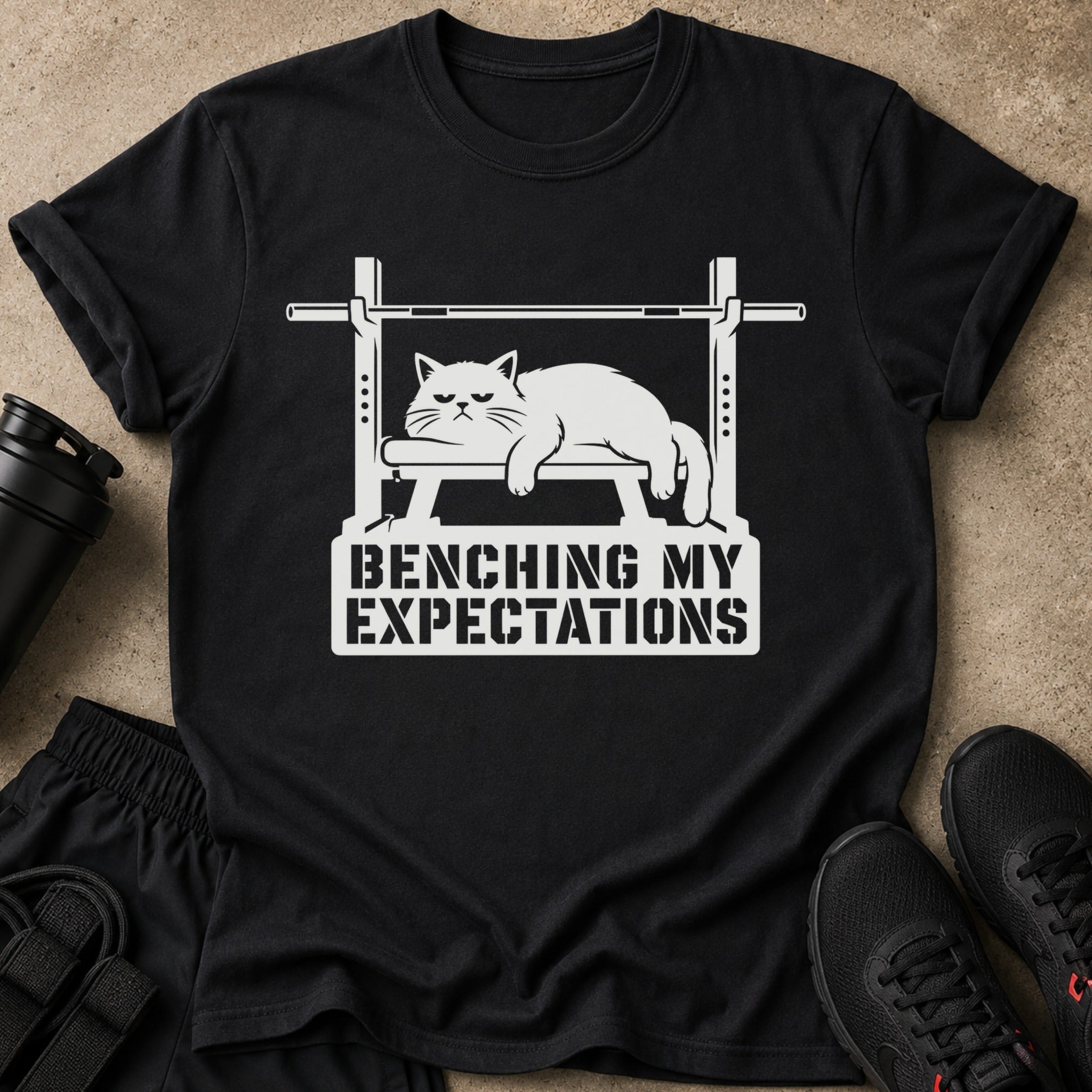 Black t-shirt with graphic of a grumpy cat on a weight bench, "BENCHING MY EXPECTATIONS" text.