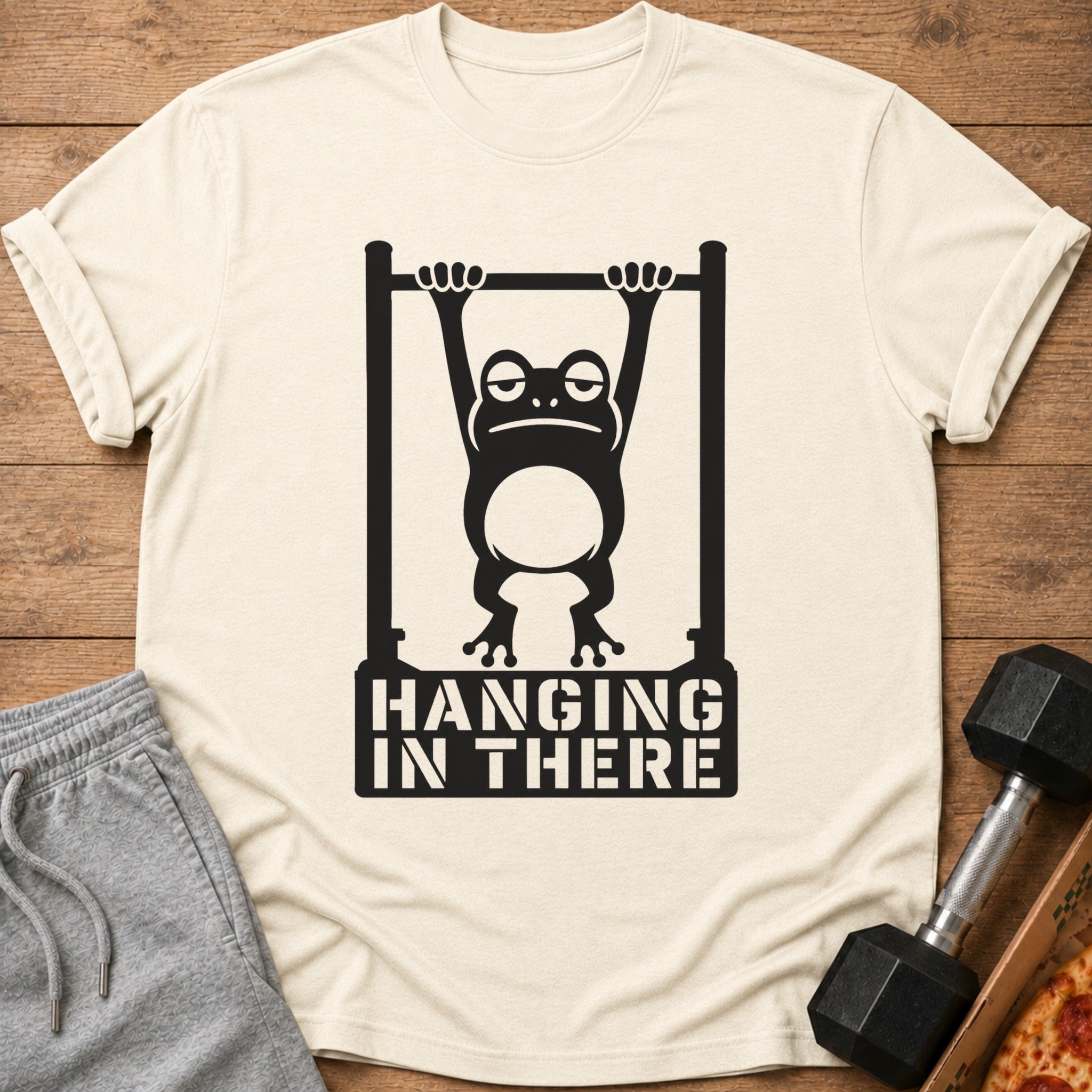 T-shirt with a black graphic of a frog hanging on a bar, saying "HANGING IN THERE".