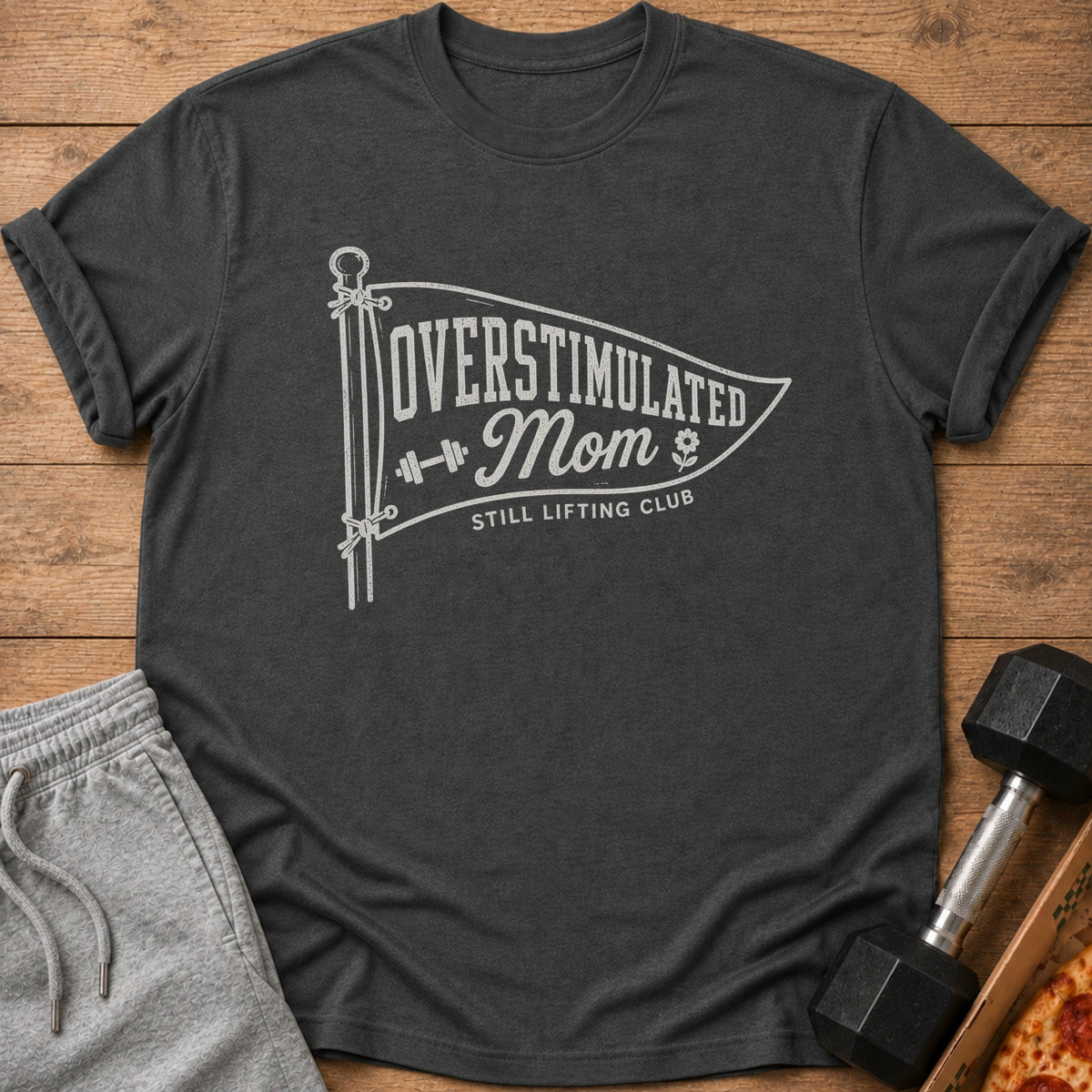 Dark gray crewneck t-shirt featuring a white pennant graphic with text reading OVERSTIMULATED Mom STILL LIFTING CLUB.