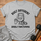 Gray t-shirt with "BUILT DIFFERENT BARELY FUNCTIONING" text and cartoon kettlebell.