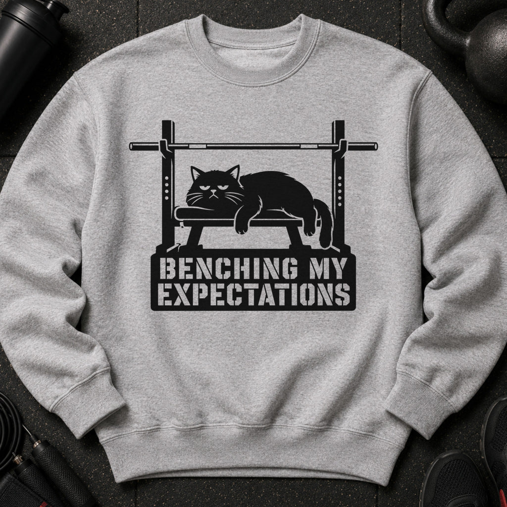 Gray sweatshirt with a black graphic of a cat on a weight bench and text "BENCHING MY EXPECTATIONS".
