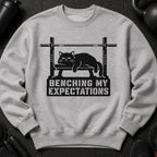 Gray sweatshirt with a black graphic of a cat on a weight bench and text "BENCHING MY EXPECTATIONS".