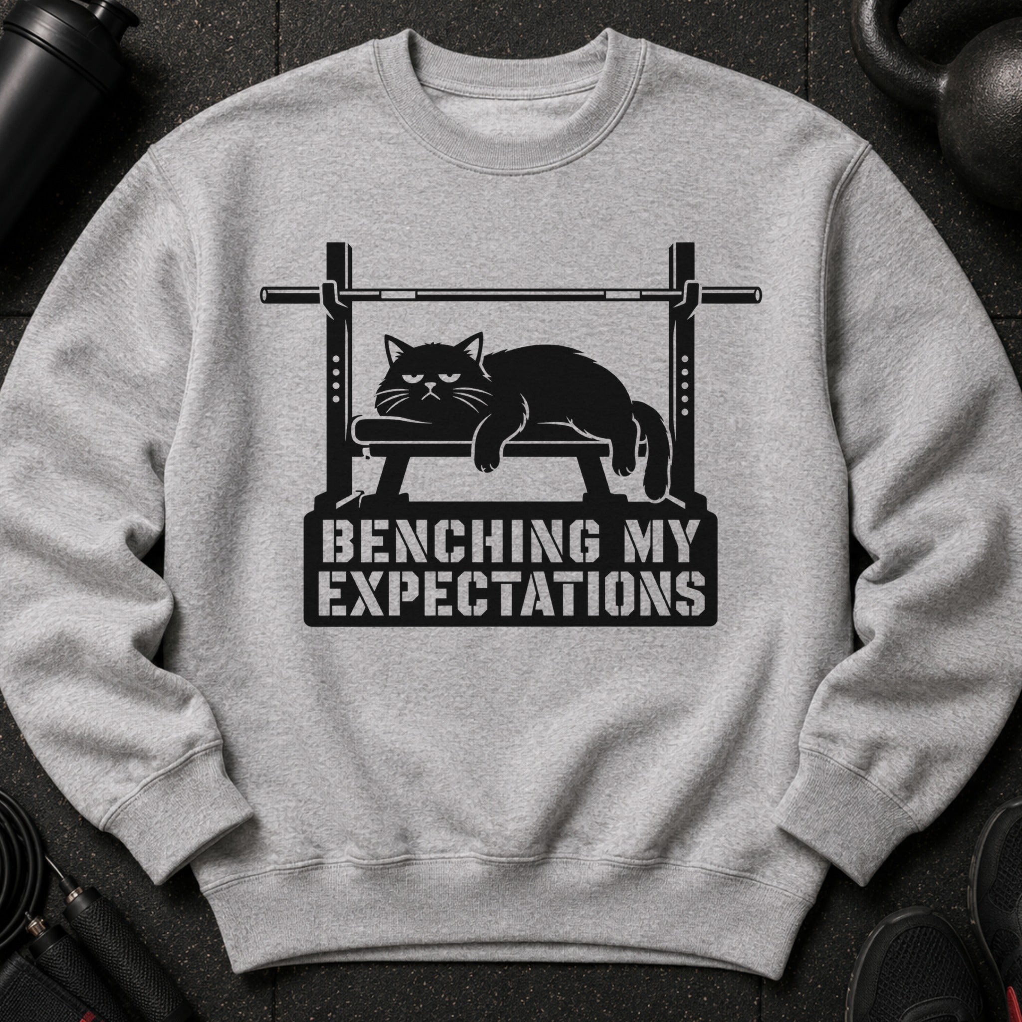Gray sweatshirt with a black graphic of a cat on a weight bench and text "BENCHING MY EXPECTATIONS".