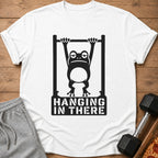 White t-shirt with a graphic of a frog hanging from a bar with the text "HANGING IN THERE".