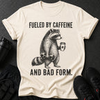 "Fueled By Caffeine And Bad Form." text on a cream t-shirt with a raccoon graphic.