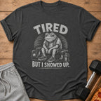 Dark grey t-shirt with a distressed graphic of a tired frog on a bench, with text "TIRED BUT I SHOWED UP."